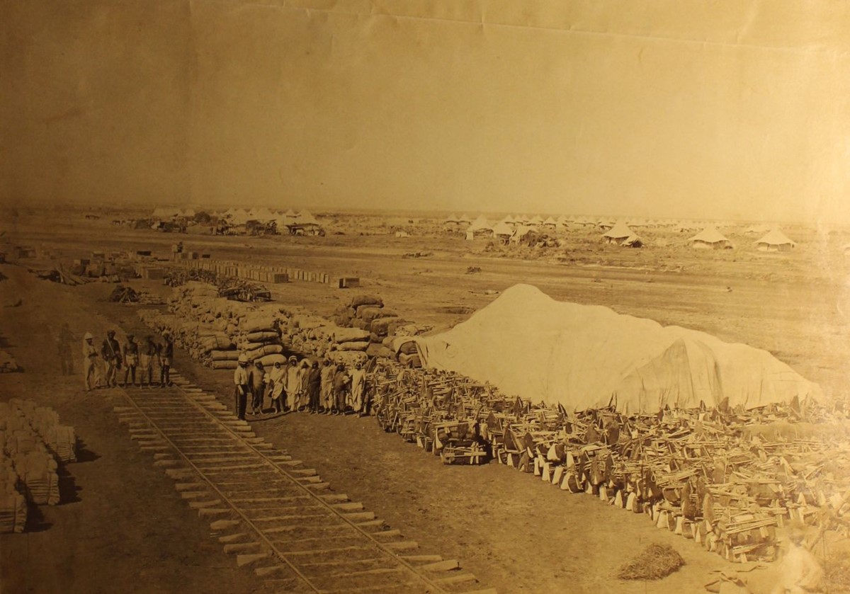 Sepia photo of a military camp with white tents, piles of supplies, and part of a roughly built railway track..