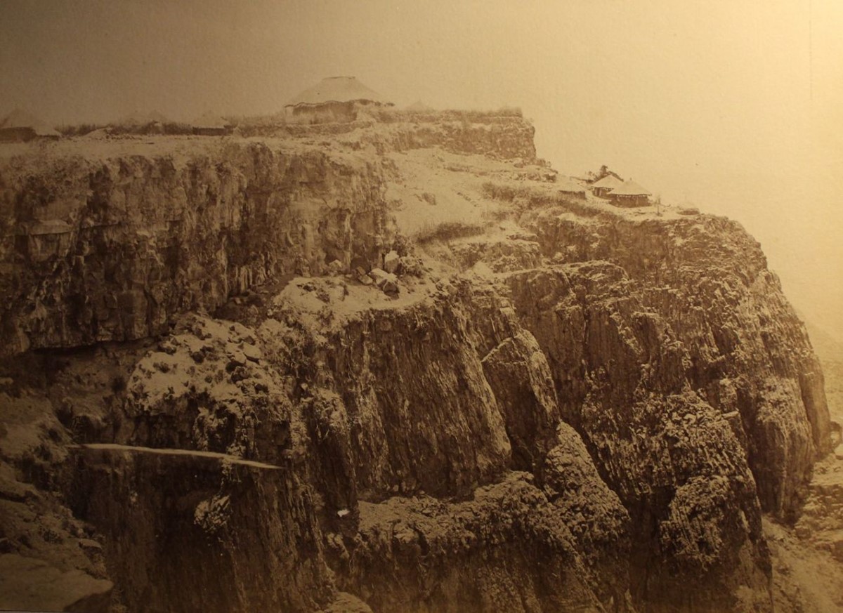photo of a large round building at the top of a plateau, with several smaller ones on a ledge below.