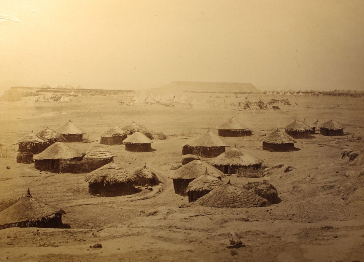 photo of a group of round buildings with thatched roofs. In the background is a group of tents.