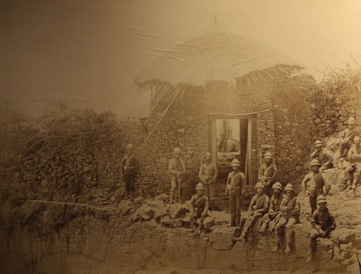 photo of soldiers in front of a stone gatehouse with a thatched roof at the top of a sheer cliff.