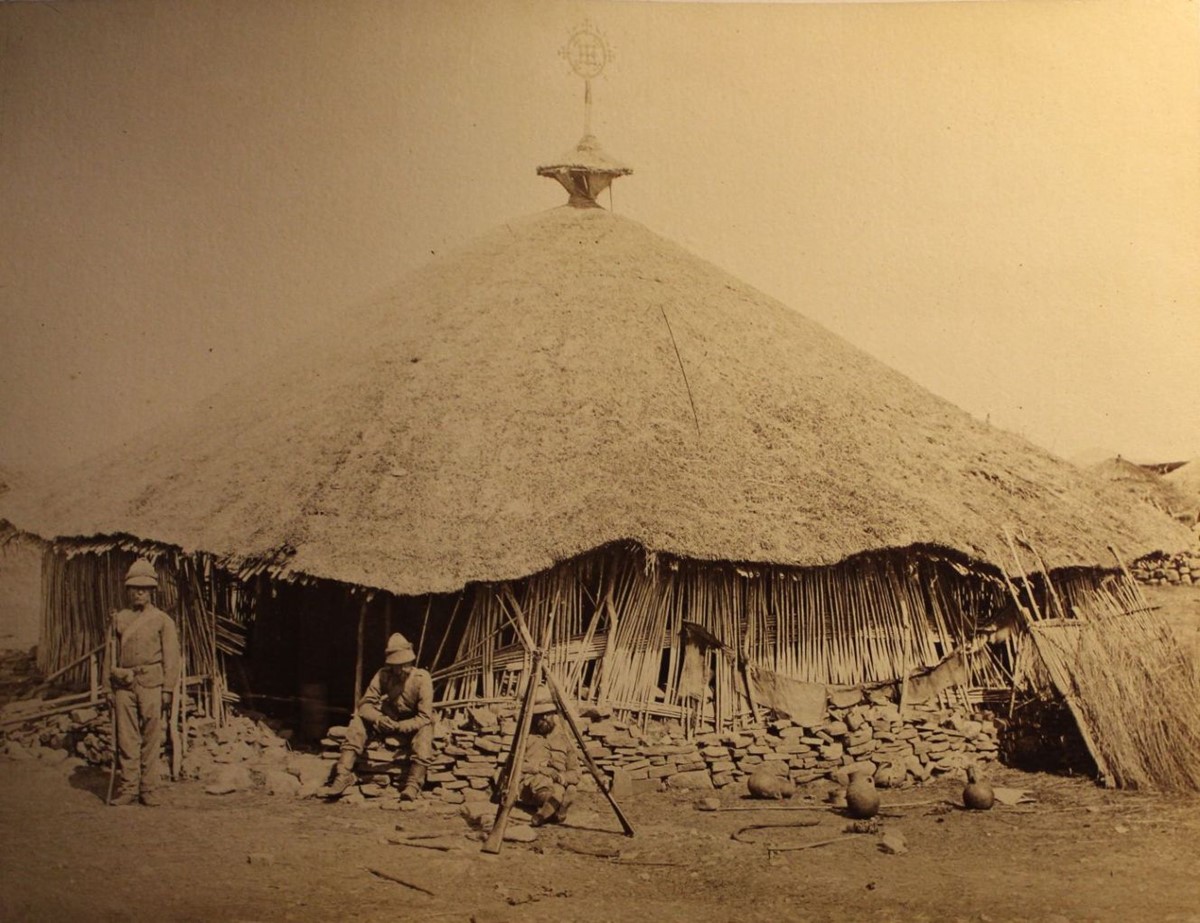 photo of a round church with an elaborate metal cross at the top of a conical thatched roof. Two soldiers are in front of it, one sitting and one standing at attention.