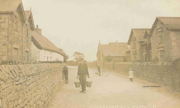 The black and white photo shows a road with walls and then houses on both sides. There is a man holding two buckets and children in the road, one boy has a bicycle. The photo dates from 1918, so the man wears a cap and the little girl has a white pinafore on.