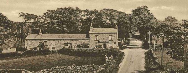 Black and white photo of a long stone building with a sign reading 'Temperance Hotel'. It stands in front of trees and beside a country lane lined with dry stone walls.