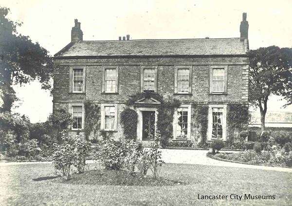 Black and white photo of Scale Hall, a two storey stone house with a symmetrical frontage. It's flanked by trees and in front of the house is a gravel carriage drive curving around a lawn and flower bed.