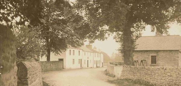 Photo looking along a road winding between stone walls, mature trees and small stone houses.