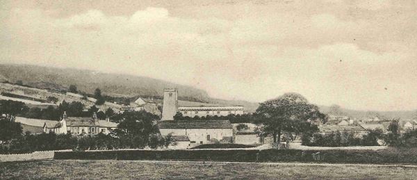 Photo of the village of Warton. Small houses and a few trees surround a stone church with a square tower. A hill rises steeply in the background.