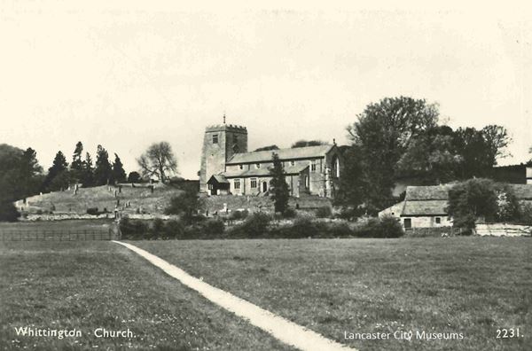 Black and white photo centred on a small stone church with a squat square tower. It sits on a low hill, surrounded by greenery and some small buildings.