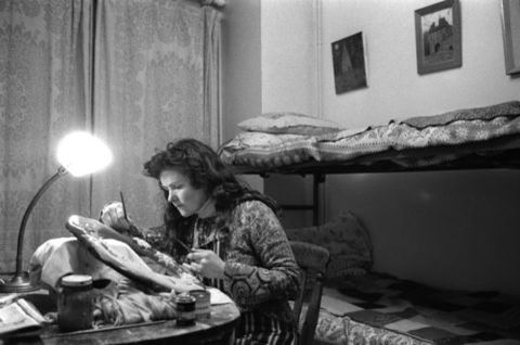Photo of Mabel Packenham-Walsh working on a sculpture. She's sitting at a desk with a bright lamp. Behind her is a set of bunk beds against the wall.