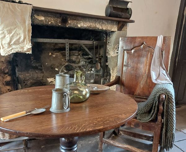 Photo of the Cottage Museum parlour. There's a wooden table and chair with a shawl draped over the back, in front of a fireplace.