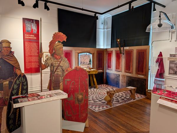 Photo of an exhibition including life sized wooden figures of roman soldiers and a corner of a room with a roman style floor mosaic, as well as cases of objects and information panels.
