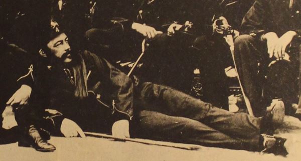 detail from a group portrait photo showing a bearded man in officer's uniform reclining on one elbow at the front of the group.